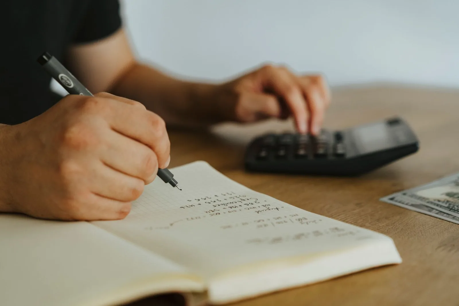 A hand writing calculations in a notebook beside a calculator on a table