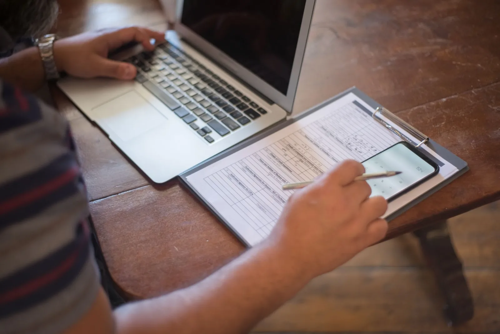 A person reviewing paper documents beside a laptop and phone on a wooden table